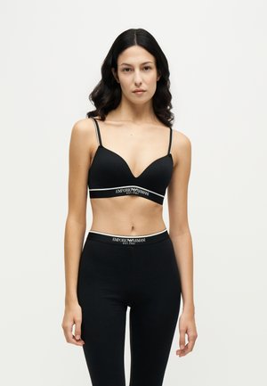 Γυναίκα με μαύρο σουτιέν και κολάν Emporio Armani με λευκές λωρίδες λογότυπου, στέκεται μπροστά σε ένα απλό φωτεινό φόντο.