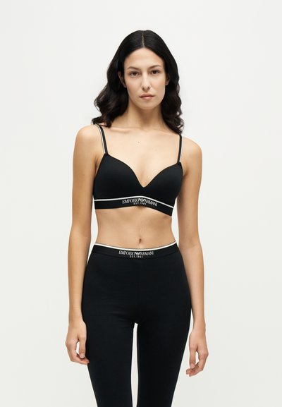 Жена, облечена в черен сутиен и клинове Emporio Armani с бели ленти с лого, стои срещу обикновен светъл фон.