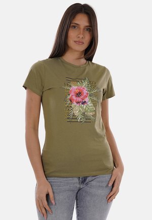 Donna che indossa una maglietta verde oliva con stampa floreale rosa e rossa e jeans azzurri, in piedi contro uno sfondo semplice.