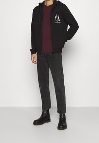 Felpa nera con zip e logo, t-shirt bordeaux, jeans grigi e stivaletti neri alla caviglia. Materiale liscio e design aderente.