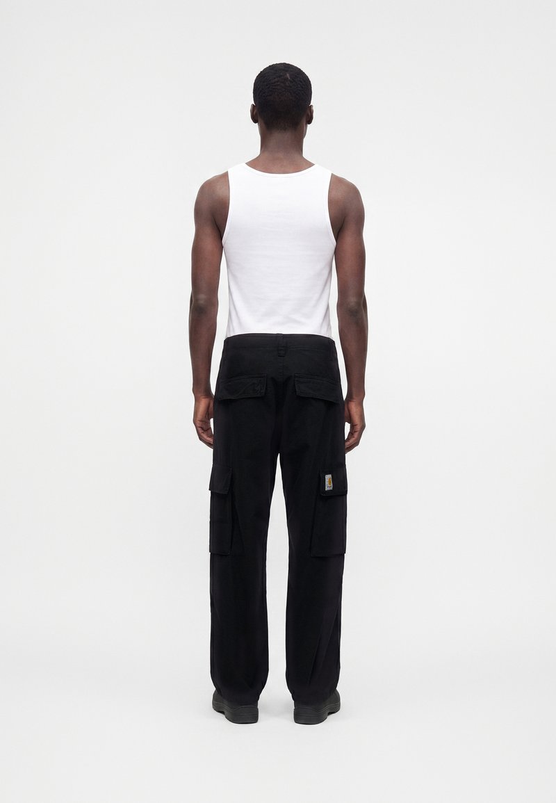 Homme debout, de dos, portant un haut sans manches blanc et un pantalon cargo noir avec plusieurs poches sur un fond uni.