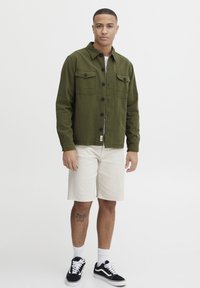 Olive green, button-up shirt jacket in cotton fabric with two chest pockets and black buttons, paired with cream shorts and black sneakers.
