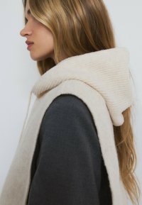 Beige gestrickte Weste mit einer gerippten Kapuze und einer weichen Textur. Getragen über einem dunkelgrauen Langarmshirt, Seitenansicht.