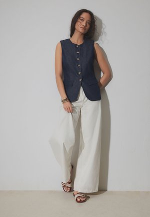 Giovane donna appoggiata al muro con gilet senza maniche blu scuro con bottoni a forma di stella, pantaloni bianchi larghi, sandali e braccialetti.