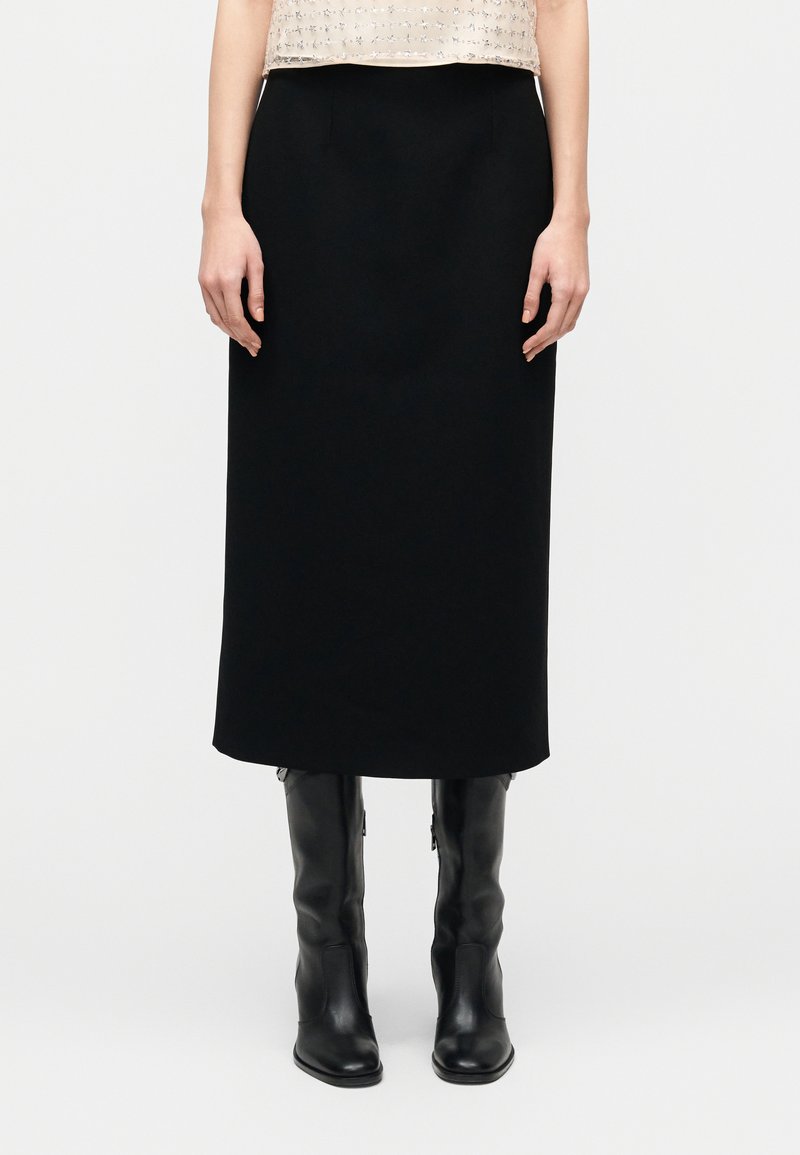 Saia midi preta feita de tecido liso, com corte direito e bolsos laterais ocultos. Combinada com botas pretas até ao joelho.