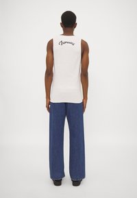 Camisa blanca sin mangas con el logo bordado en negro "Fiorucci" en la parte superior de la espalda, combinada con pantalones de mezclilla azules sueltos.