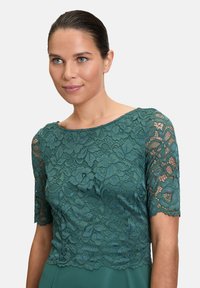 Robe verte avec un corsage en dentelle ornée de motifs floraux et de manches courtes, tissu lisse en dessous, encolure ronde et ourlet festonné.