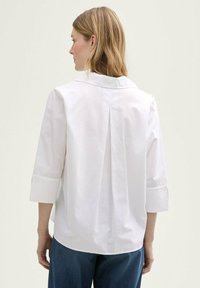 Chemise en coton blanc avec col et manches trois-quarts, présentant un pli central au dos et un ourlet courbé. Design et texture minimalistes.