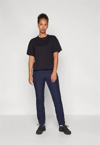 Νεαρή γυναίκα στέκεται, φορώντας μαύρη αέρινη μπλούζα με κοντά μανίκια, ναυτικές παντελόνες με φερμουάρ και μαύρα αθλητικά παπούτσια μπροστά σε απλό φόντο.