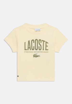 Κοντομάνικη μπλούζα εκρού με το κείμενο "LACOSTE FRENCH SPORTSWEAR" και πράσινο λογότυπο κροκόδειλου μπροστά.