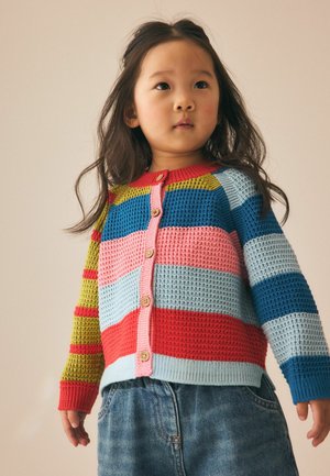 Young child with long dark hair wearing a colorful striped knit cardigan and blue denim jeans looking slightly upward.