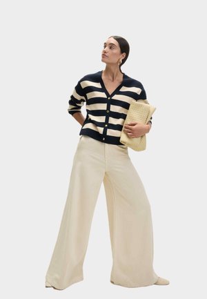 Navyblauwe en crème gestreepte cardigan met knopen, gecombineerd met brede crèmekleurige broeken. Accessoire: gestructureerde beige clutch. Eenvoudig, modern ontwerp.