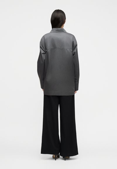 Γυναίκα που στέκεται με την πλάτη στο φακό φορώντας γκρι φαρδιά μπουφάν, μαύρο παντελόνι με φαρδιά μπατζάκια και μαύρες ψηλοτάκουνες γόβες, σε λευκό φόντο.