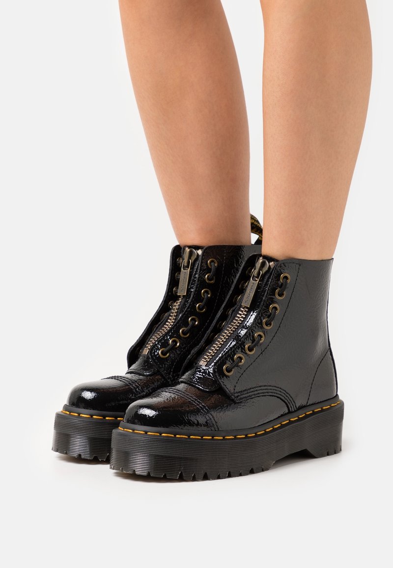 Bottes en cuir noir brillant à bout rond avec semelles épaisses, fermetures éclair à l'avant et coutures jaunes portées sur des jambes nues sur fond blanc.