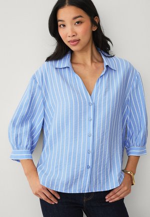 Junge Frau posiert in einer locker sitzenden hellblauen Bluse mit vertikalen weißen Streifen und dreiviertellangen, aufgebauschten Ärmeln, kombiniert mit dunklen Jeans.