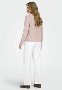 Lys rosa strikket genser med brodert blomstermotiv på venstre erme, sammen med hvite vide bukser og brune sko.