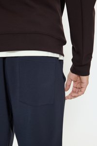 Osoba ubrana w ciemnogranatowe spodnie z tylną kieszenią i ciemny, długorękawowy top, z ręką spoczywającą obok ciała na jasnym tle.