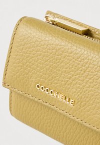 Primer plano de una billetera de cuero amarillo texturizado con cremallera dorada y el logo de Coccinelle en relieve en la solapa delantera.