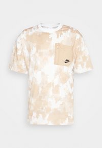 Beige och vit mönstrad t-shirt med prickdesign, med en framficka med dragkedja och en svart Nike-logotyp på fickan.