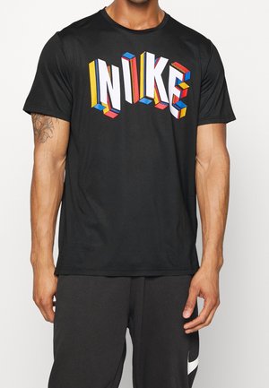Hombre con camiseta negra de Nike con letras tridimensionales de colores que forman "NIKE" y pantalones negros con logo blanco en la pierna izquierda.