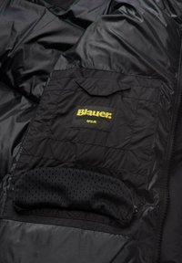Giacca a vento nera con una grande tasca a toppa con "Blauer" e "USA" in giallo, e dettaglio della tasca con zip foderata in rete.