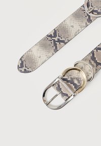 Ζώνη από ύφασμα με σχέδιο φολίδας φιδιού σε αποχρώσεις μπεζ και γκρι. Διαθέτει αγκράφα από ασημένιο μέταλλο με χρυσή λεπτομέρεια. Λεία υφή.