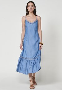 Robe maxi en denim bleu clair avec des bretelles spaghetti réglables, décolleté en V et bordure brodée à l'ourlet. Texture douce avec un bas évasé.