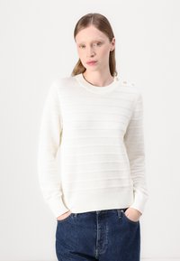Hvit strikkegenser med horisontale striper, ribbestrikkede mansjetter og utringning. Har gullknapper på skulderen. Kombinert med mørkeblå jeans.