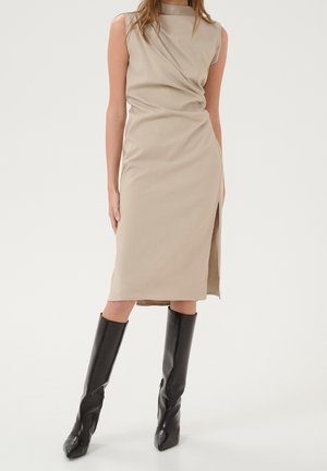 Femme portant une robe midi beige sans manches avec une fente latérale et des bottes noires hautes à talons, debout devant un fond uni.