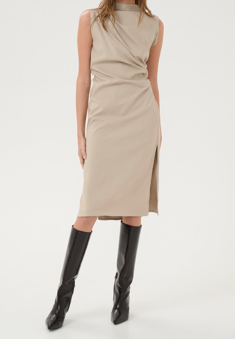 Femme portant une robe midi beige sans manches avec une fente latérale et des bottes noires hautes à talons, debout devant un fond uni.