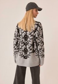 El suéter presenta una base gris con un patrón floral negro, puños y dobladillo acanalados. Combinado con pantalones gris oscuro y una gorra a juego.