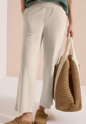 Pantaloni beige a gamba larga con vita elastica, scarpe marroni senza lacci e una mano che tiene una borsa intrecciata con manici chiari.