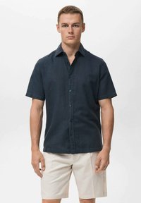 Camisa de botones de manga corta en un oscuro tono navy, hecha de una tela ligera, con un cuello puntiagudo y un dobladillo recto, combinada con pantalones cortos beige.