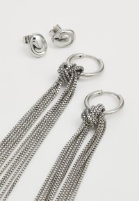Cercei din metal de nuanță argintie, cu două designuri: lanțuri în stil nod cu multiple fire și cercei studs ovali netezi.