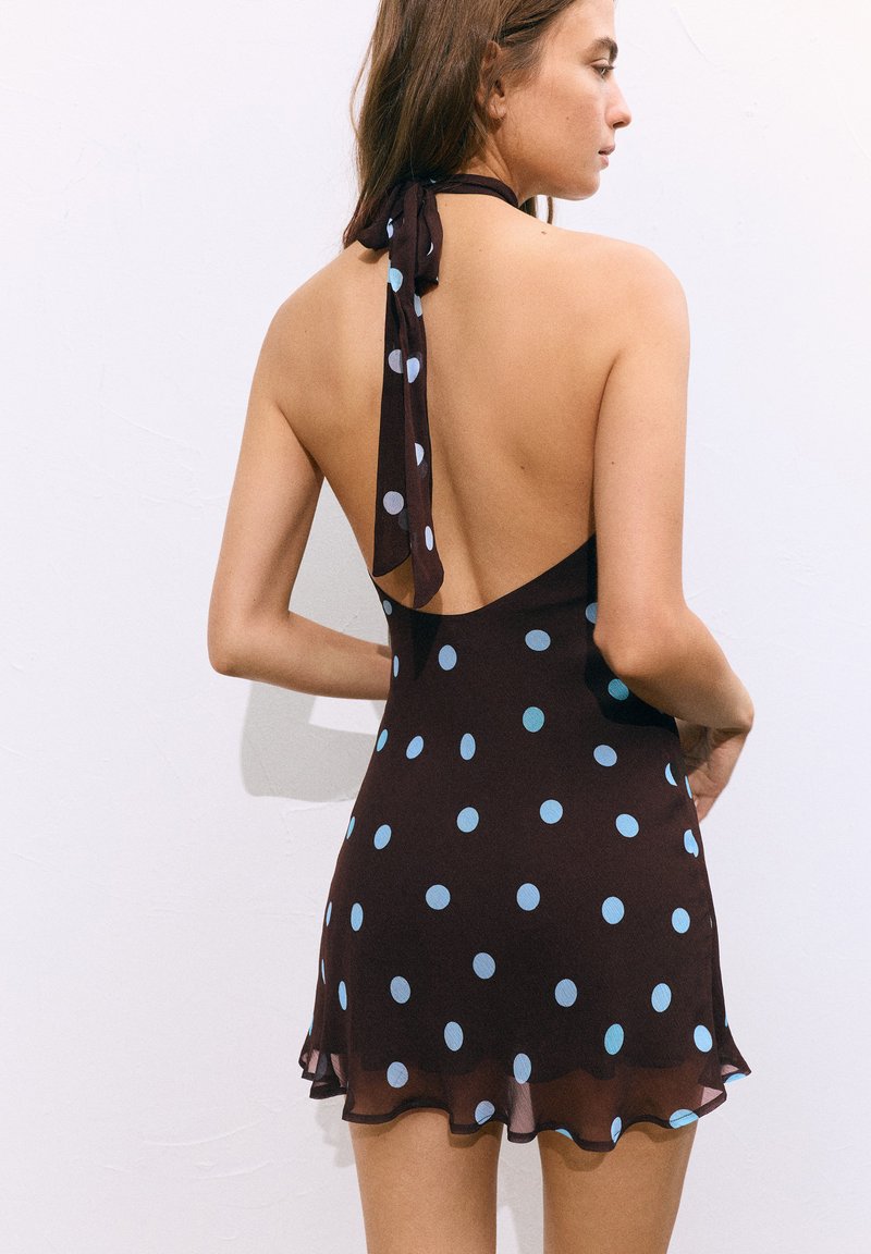 Femme portant une robe noire à col halter avec des pois bleus, montrant un dos décolleté et un ruban noué au cou.