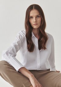 Giovane donna con lunghi capelli castani, che indossa una camicia bianca a bottoni e pantaloni beige, seduta a gambe incrociate su uno sfondo neutro.