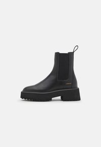 Musta nahkainen nilkkasaapas, jossa on joustavat sivupaneelit, paksu korokepohja ja takana vetolenkki. Kultainen logo sivulla.