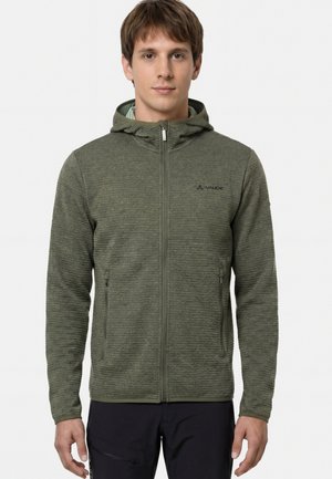 Giovane uomo che indossa una felpa con cappuccio verde oliva testurizzata con cerniera e un piccolo logo sul petto, pantaloni neri con cerniera, in piedi su sfondo bianco.