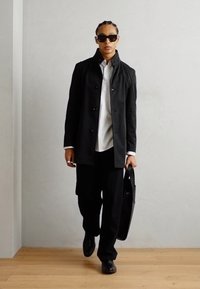 Zwarte overjas met knopen, witte shirt en zwarte broek. Accessoires zijn zwarte zonnebril en een zwarte handtas, met een achtergrond van een houten vloer met textuur.