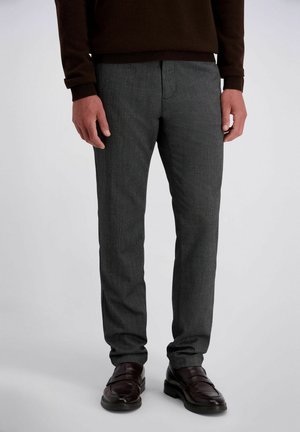 Man draagt donkergrijze pantalon, donkerbruine trui en donkerbruine leren schoenen, staand tegen een effen lichte achtergrond.