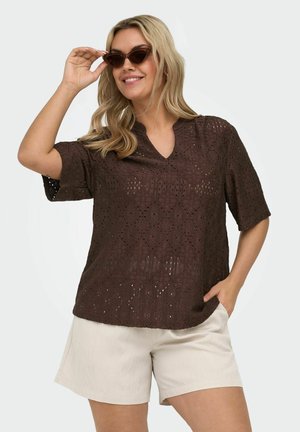 Kvinne iført brun kortermet blonder topp, beige shorts og katteøye-solbriller, smilende med én hånd som justerer solbrillene.