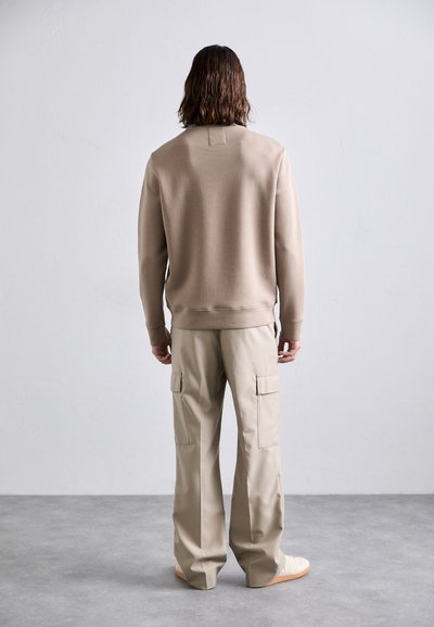 Person with shoulder-length hair wearing a beige sweatshirt and light tan cargo pants, standing on a gray floor against a plain white wall.