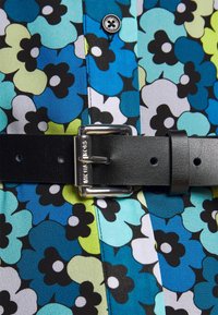 Tissu à motif floral avec des fleurs bleues, vertes et noires. Une ceinture en cuir noir avec une boucle en argent se resserre à la taille.