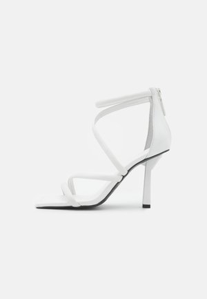 Sandalo bianco con tacco alto, dotato di tre sottili cinturini sulla parte anteriore del piede e di un cinturino alla caviglia con cerniera sul retro, su uno sfondo grigio chiaro uniforme.