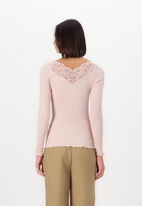 Langlijf pink geribbelde top met een geschulpte zoom en kantafwerking bij de achterkant van de hals, gecombineerd met beige broek.