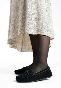 Zwarte suede moccasin-stijl pantoffels met een ronde neus, decoratieve strik en geplooide textuur. Draag ze met een lichte gespikkelde midi-jurk en zwarte maillot.