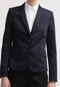 Persona que lleva puesta una chaqueta negra ajustada con dos botones sobre una camisa blanca con cuello, combinada con pantalones negros.