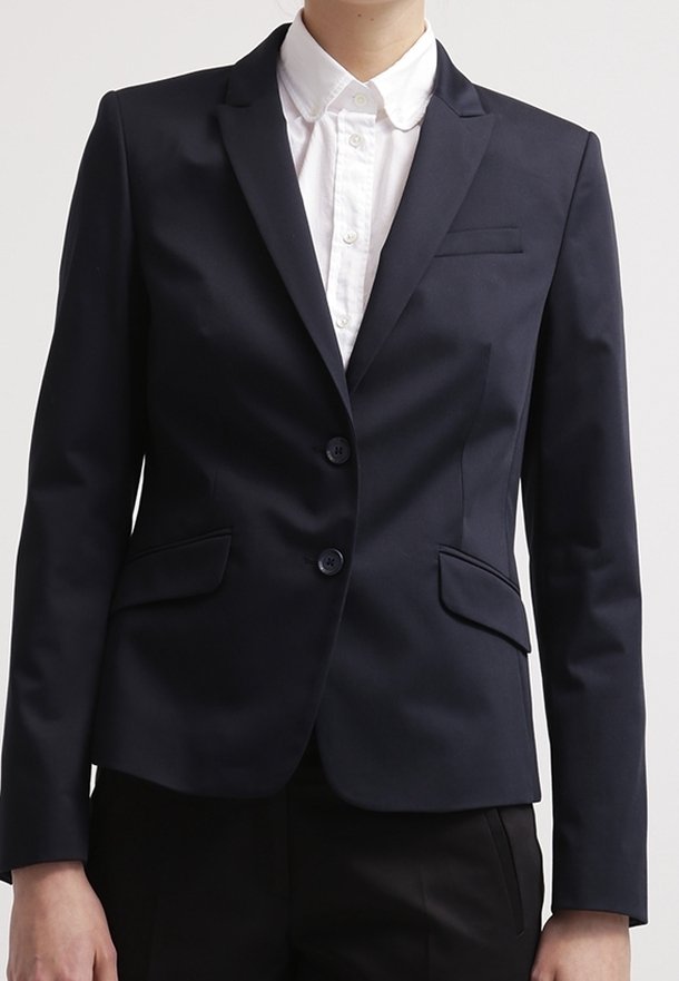 Persona que lleva puesta una chaqueta negra ajustada con dos botones sobre una camisa blanca con cuello, combinada con pantalones negros.