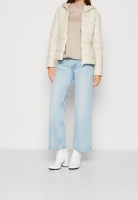 Lys beige pufferjakke med hætte, parret med en beige top og lyseblå vide ben jeans. Hvide hælede ankelstøvler med en sort accent.