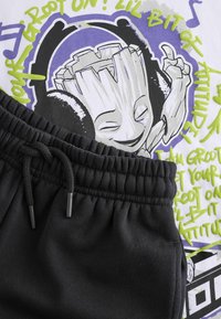 Bavlněné grafické tričko s potiskem kreslené postavičky v sluchátkách, bílé barvy, s fialovými a zelenými akcenty. Doplňují je černé tepláky se stahovací šňůrkou.